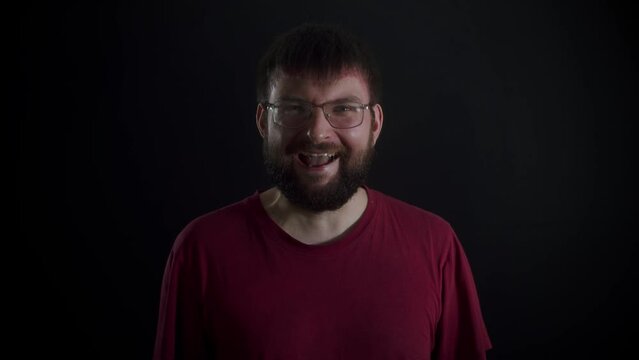 A bearded man wearing glasses and a burgundy T-shirt laughing, covering his mouth with his hand against a black monochromatic background. Front view, slowmo.