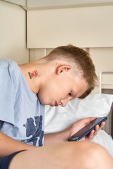 Naklejka premium Teen boy with short blond hair reads electronic book with smartphone sitting on upper rack traveling in train car closeup