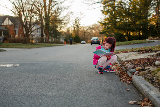 Little girl squats down in street to write notes in journal at sunset