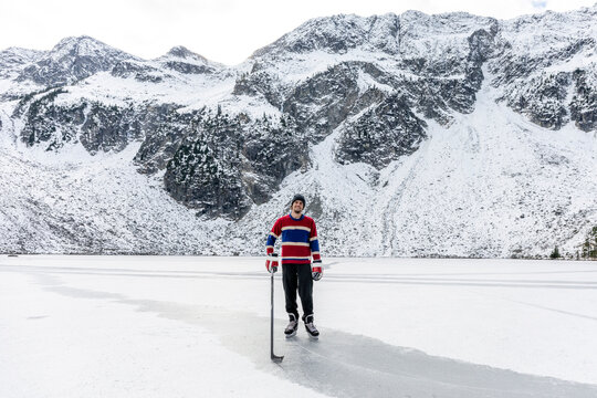 Full Body Young Man With Stick And Puck Smiling And Looking At Camera While Standing On Ice Of Frozen Lake Near Snowy Mountain During Hockey Game In Countryside Of British Columbia, Canada