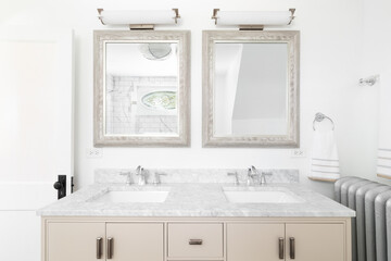 A bathroom with a cream colored double vanity cabinet, marble countertop, and lights hanging above...