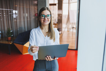 Cheerful young fair-haired business woman of European appearance in a white shirt and glasses holds a portable laptop in her hands, stands near a large window in the office on the top floor © DSMT