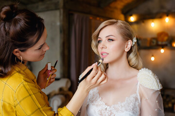 A makeup artist paints the lips of a young blonde girl with lipstick on her wedding day. Selective focus. Morning of the Bride