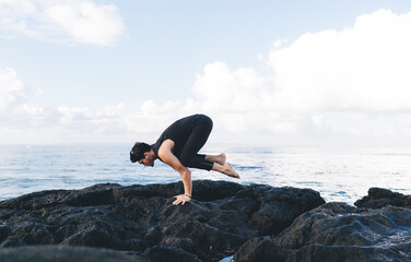 Side vie of strong male yogi doing balance hands stand during morning time for intensive practice at coastline, Caucaisan man in black sportive wear breathing while exercising at seashore