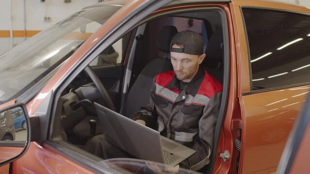 Mechanic sitting in car and working on laptop while doing vehicle diagnostic test in auto service garage