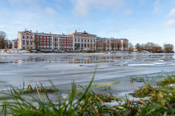 Fototapeta na wymiar Low angle view to baroque house on the river in winter. Palace in the city of Jelgava, Latvia, Europe. Historical red-white residential building on the frozen river bank.