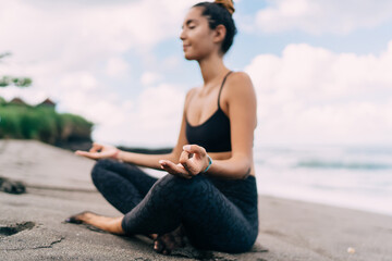 Fototapeta na wymiar Harmony and meditation training during morning yoga at coastline beach, blurred female in sportswear sitting in lotus pose during aerobic pilates for exercising own body concentration in asana