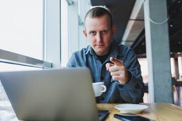 Fototapeta premium Successful happy business man is sitting at a table in a cafe, holding a cup of coffee and using a laptop.