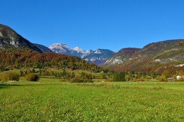 View of Tosc, Ablanca and Draski vrh mountain peaks in Julian alps and Triglav national park, Slovenia in autumn with the hills covered in red colored forest and a meadow in front