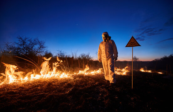 Firefighter working in field with wildfire at night. Man in suit and gas mask near burning grass with smoke and yellow triangle with skull and crossbones warning sign. Natural disaster concept.