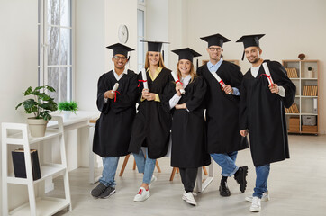 Portrait of smiling multiethnic college graduates in mantles and hats pose in classroom with...