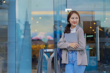 Asian woman holding a tablet with a bag standing in front of a shopping mall selling work-studying ideas outdoors.