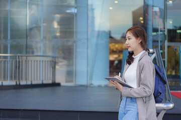 Asian woman holding a tablet with a bag standing in front of a shopping mall selling work-studying ideas outdoors.