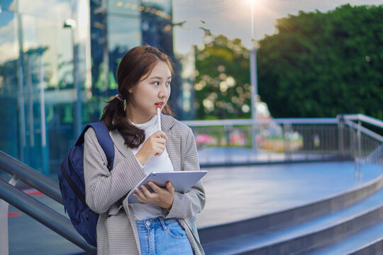 Asian woman holding a tablet with a bag standing in front of a shopping mall selling work-studying ideas outdoors.