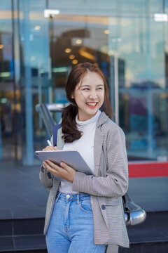 Asian woman holding a tablet with a bag standing in front of a shopping mall selling work-studying ideas outdoors.