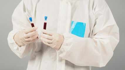 Hands are holding two blood test tubes on grey background. Hand wears a PPE suit and a white...