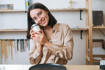 Portrait of charming smiling young woman in glasses holding little transparent glass jar with hand-made scented candle, looking at camera. Process of making handmade natural candle at workshop.