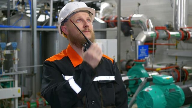 Technological engineer in a white helmet transmits instructions to the staff using a walkie-talkie. A bearded man with blue eyes at a workplace in an industrial workshop.