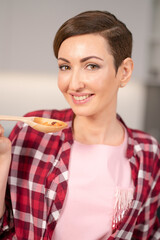 Close up housewife in red plaid shirt and short hair cooking soup on modern kitchen. Smiling woman testing her dish. Chef woman having fun at kitchen