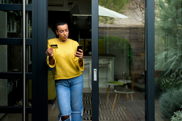 Portrait of casual relaxed woman using phone for online payment on doorstep of her home shopping online from home leaning on door frame using credit card casual entrepreneur doing business from home