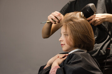 Charming girl enjoys hair care in beauty salon. Child looks in mirror and enthusiastically presses her hands to her chest while hairdresser is styling her hair. Short bob haircut