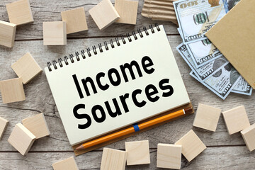 INCOME SOURCES text on white notebook pages near wood blocks. there are a lot of dollar bills on...