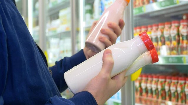 A man standing at a glass display case with dairy products chooses fruit yogurt while reading the description and ingredients In the hands of three bottles with transparent glass in which a milk drink