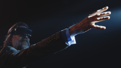 Middle aged male creator showing viewers, how augmented reality glasses work near LED screen and touching something in the air during a Metaverse event in a dark lit room with spotlights