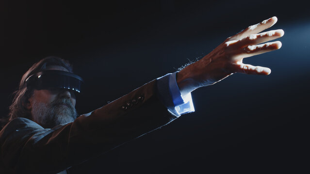 Middle aged male creator showing viewers, how augmented reality glasses work near LED screen and touching something in the air during a Metaverse event in a dark lit room with spotlights