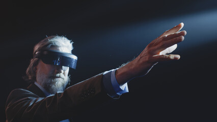 Middle aged male creator showing viewers, how augmented reality glasses work near LED screen and touching something in the air during a Metaverse event in a dark lit room with spotlights