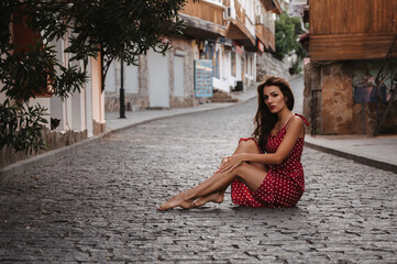Fototapeta na wymiar Sexy young Caucasian girl in a red dress posing sitting on a paving stone on a beautiful European street in Italy