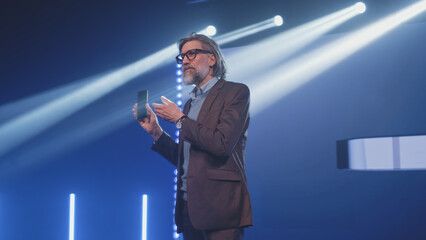 A male speaker on stage holding a new smartphone and talking about the characteristics, while...
