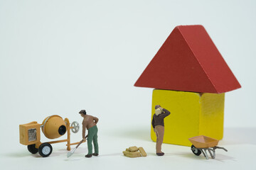 A team of workers with a cement mixer and a wheelbarrow works on a yellow house with a red roof,...