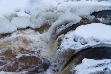 Landscape photo of non-freezing running river with fast current and rapids on its way flowing between ice-covered white banks against backdrop of snowy forest in distance on gloomy cold winter day