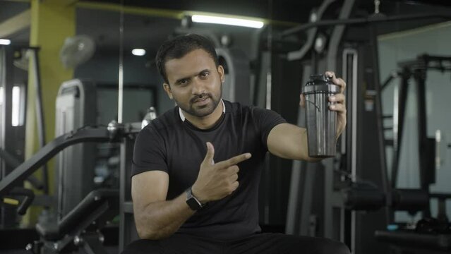young indian muscle builder with protein powder on shaker bottle showing to camera at gym - concept of advertisment, muscle building and nutrition supplement promotion.