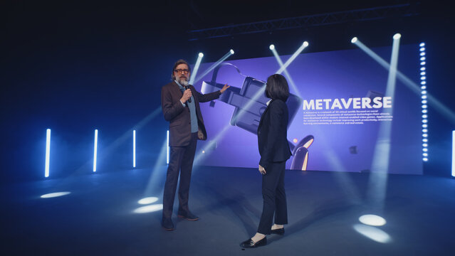 A female presenter interviewing a male developer of a new augmented reality device during the presentation of meta universe in a modern studio with spotlights, near the LED screen in 3D objects