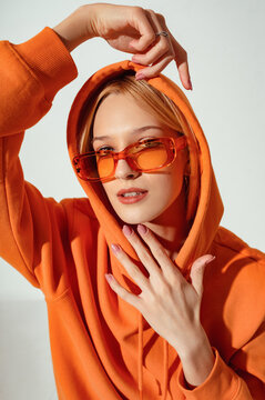 Fashion portrait of confident woman wearing trendy orange color sunglasses, oversized hoodie. Model looks at camera. Indoor, studio close up fashion portrait