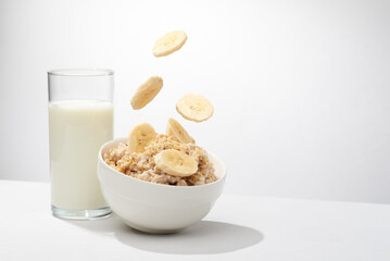 Oat porridge with levitating banana`s slices, walnut`s crumbs and glass of milk on a white background. Minimalistic photo with place for your text.