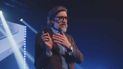 Presenter standing on stage with a new contemporary smartphone at the presentation, and explaining to the audience the new technologies of a mobile phone in front of an LED screen with a 3D mock-up - obrazy, fototapety, plakaty