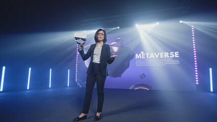 A female speaker in casual clothes in a studio with LED screen holding new virtual reality glasses at a Metaverse presentation and speaking about new modern technologies, in the virtual world