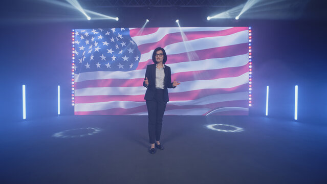 Female Presenter In Suit On Stage Talking About The Latest Political And Public News Of The United States On TV Broadcast In The Modern Studio, With Spotlights Near The American Flag On The LED Screen