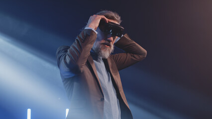 Fototapeta na wymiar Mature developer in augmented reality glasses gesturing and touching something in the air at a Metaverse event in front of an LED screen, explaining to the audience how virtual reality devices work