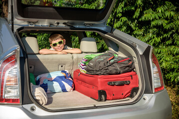 Adorable kid boy wearing sunglasses sitting in car trunk. Portrait of Happy child with open car boot while waiting for parent get ready for vocation. Family trip traveling by car concept