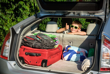 Adorable kid boy wearing sunglasses sitting in car trunk. Portrait of Happy child with open car boot while waiting for parent get ready for vocation. Family trip traveling by car concept