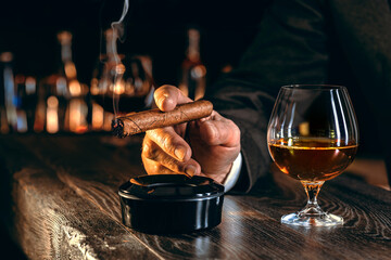 Man's hands with a cigar, elegant glass of brandy on the bar counter. Alcoholic drinks, cognac,...