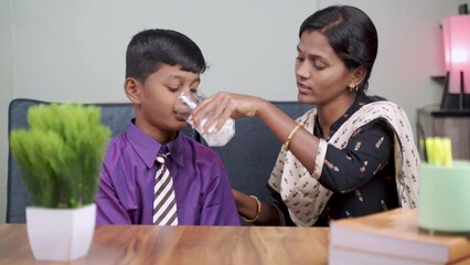 Indian mother helping school kid to drink glass of milk before going to school at home - concept of healthy childhood lifestyle, wellbeing and caring.