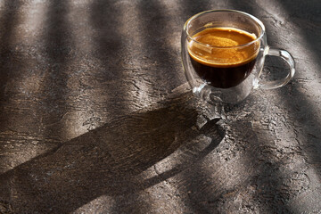 Glass cup of morning double espresso in sunlight on dark textured table, close-up, shallow depth of field