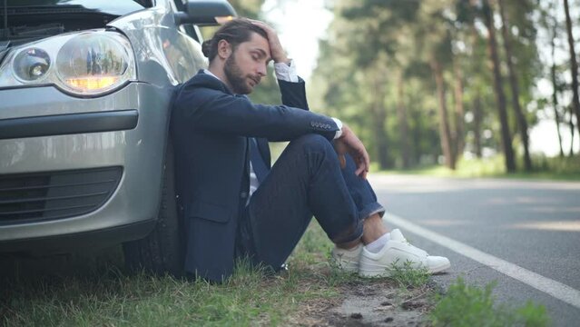Side view hopeless young man sitting at car breakdown lights on with vehicle passing on suburban road. Sad stressed Caucasian male driver outdoors on sunny day thinking. Accident concept