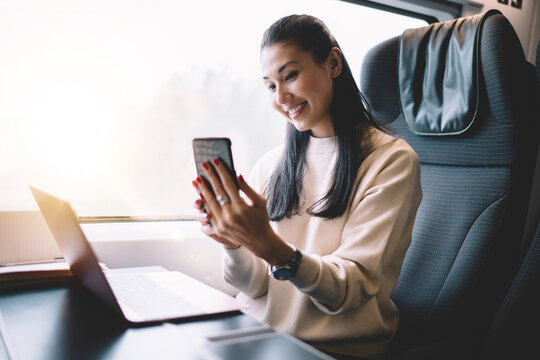 Cheerful female blogger working during trip on railroad sitting in train with wireless internet connection making picture on frontage mobile phone camera, happy woman posing for selfie on smartphone