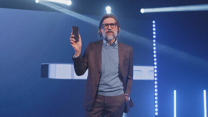 A male speaker on stage holding a new smartphone and talking about the characteristics, while standing in front of an LED screen with a phone mock-up and an inscription during the presentation